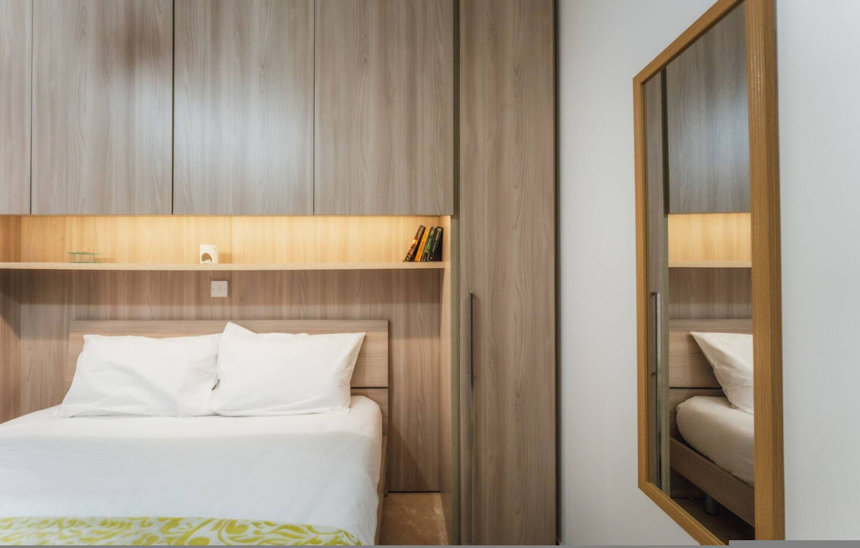 A hotel room with a bed, a shelf with books, and a mirror.