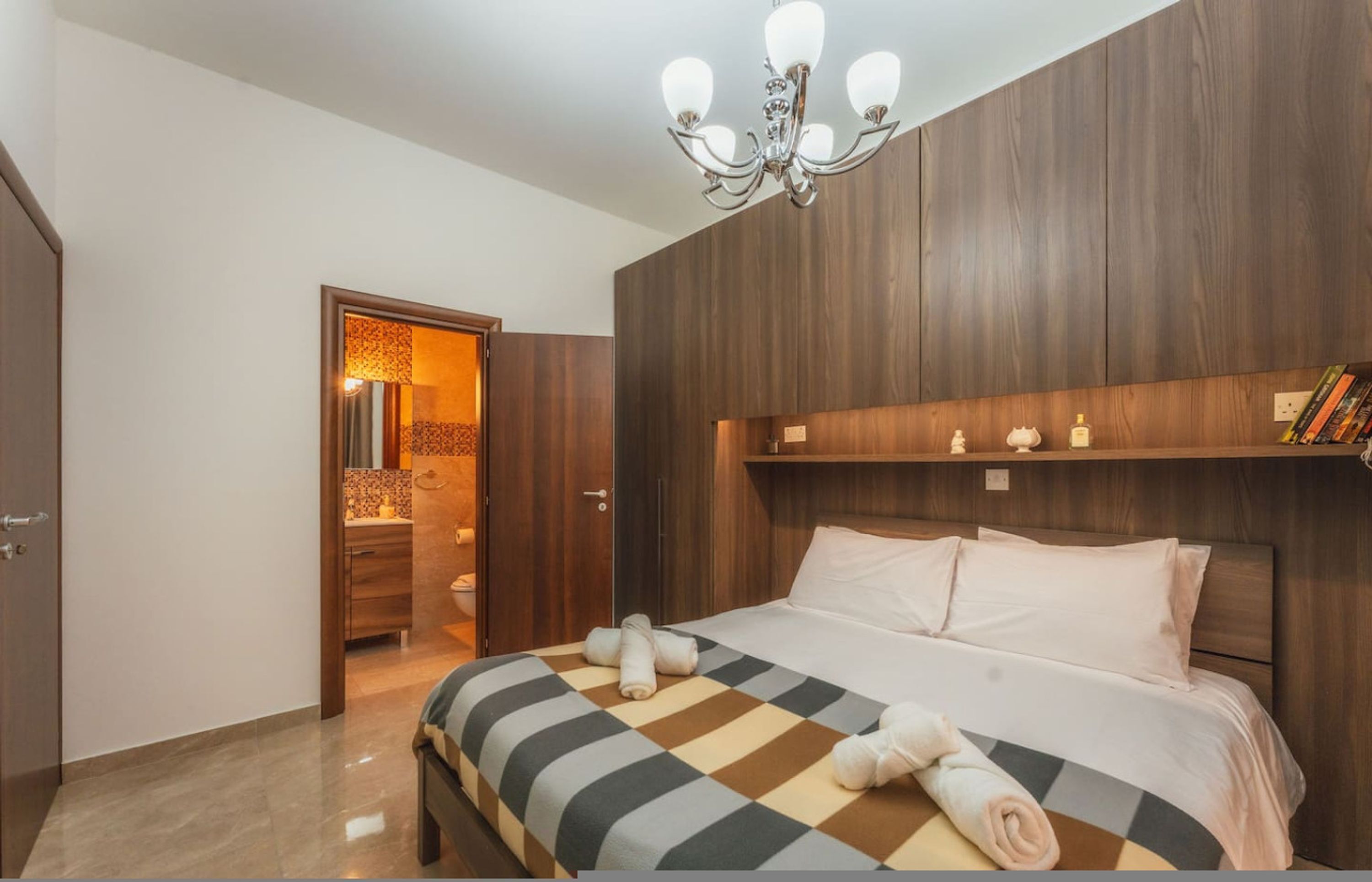 A bedroom with a bed, a chandelier, a wooden headboard, and a nightstand with books and a lamp.