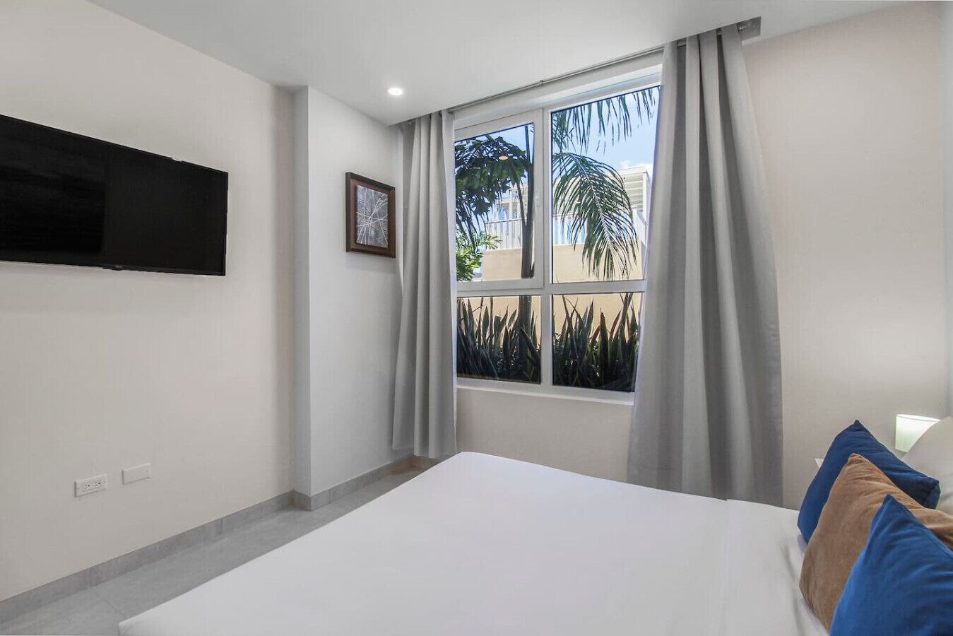 A bedroom with a bed, a TV mounted on the wall, a window with curtains, and a view of palm trees outside.