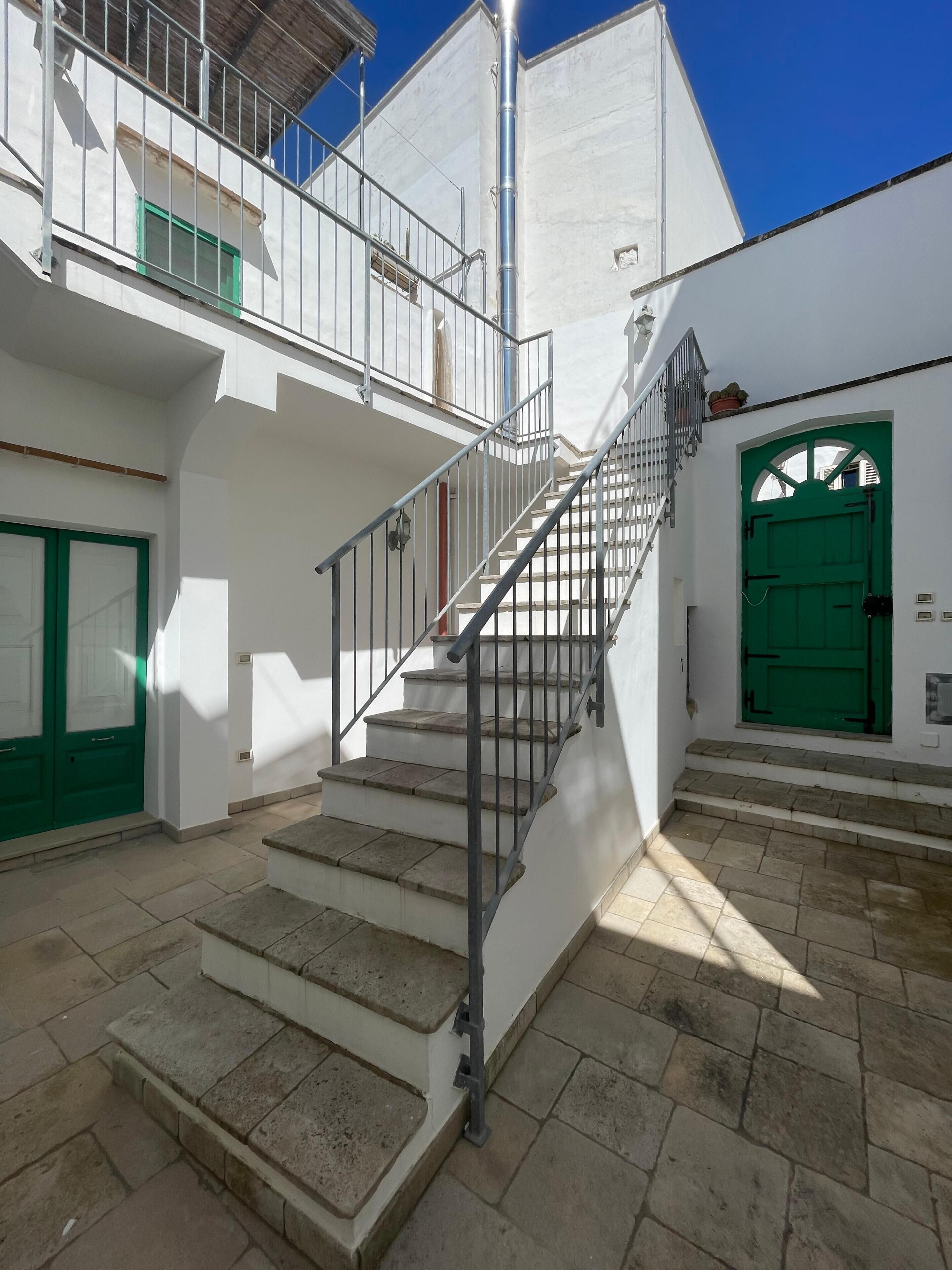 Una scala conduce a un balcone con ringhiera metallica, una porta verde e una facciata di un edificio bianco.