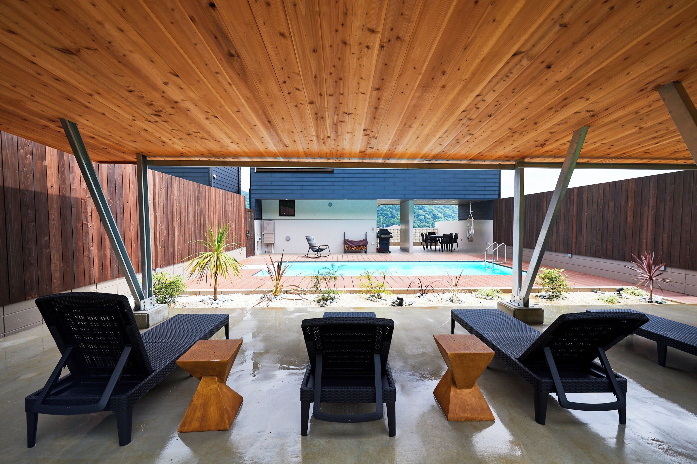 Ein Außenbereich mit einer Sitzecke, drei Liegestühlen, einem Tisch, einer Holzbalkendecke und Blick auf einen Swimmingpool.