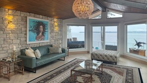 A spacious room with a stone wall, a large painting, a green sofa, a tufted ottoman, a glass coffee table, and a view of the water. - Perfect 6-bedroom house in Blaine for your next getaway (Blaine)