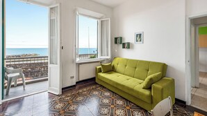 Et værelse med en grøn sofa, et mønstret gulv, havudsigt og en balkon med stole.