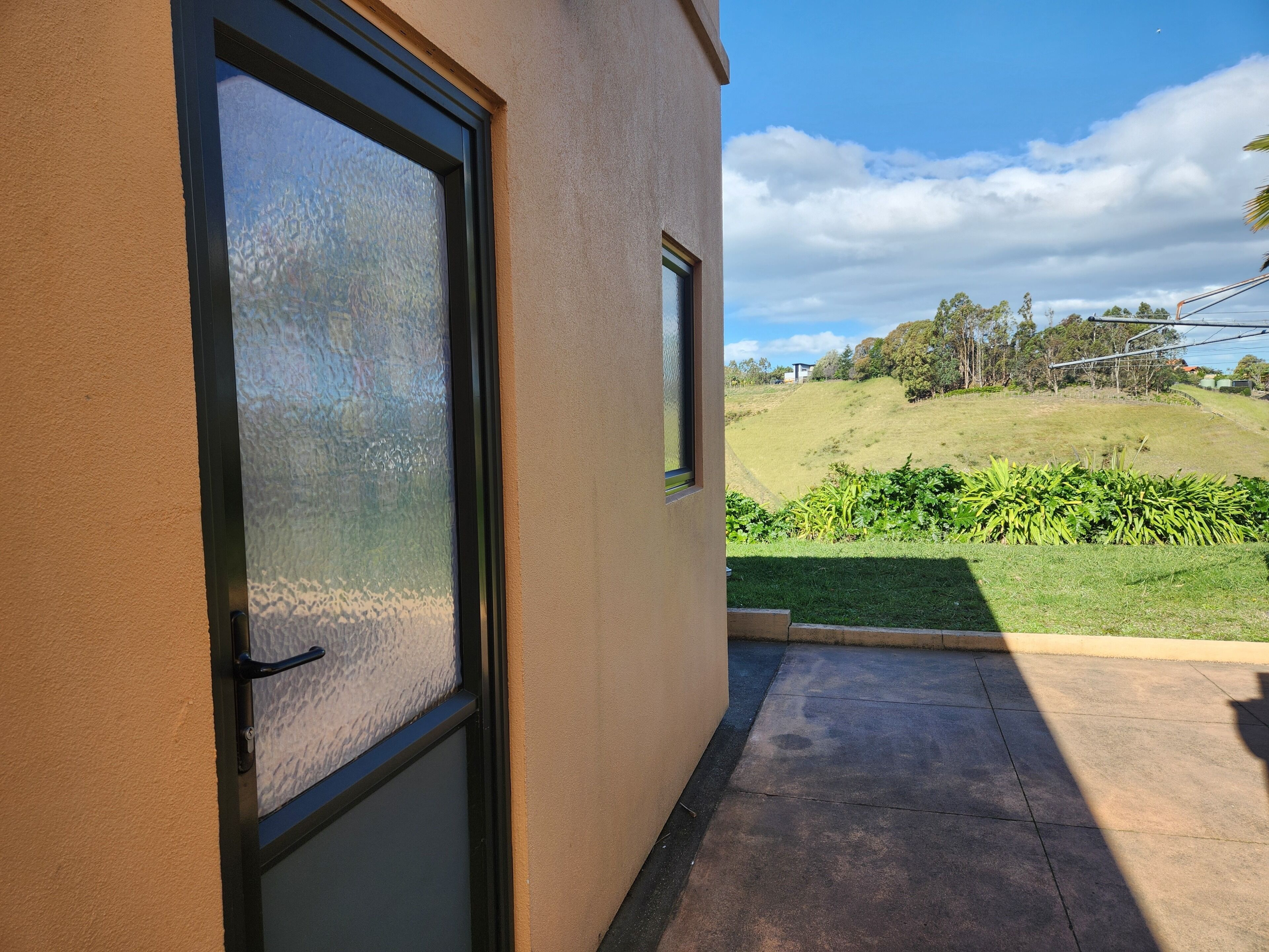 A building with a large window, a concrete patio, and a view of a grassy hill with trees.