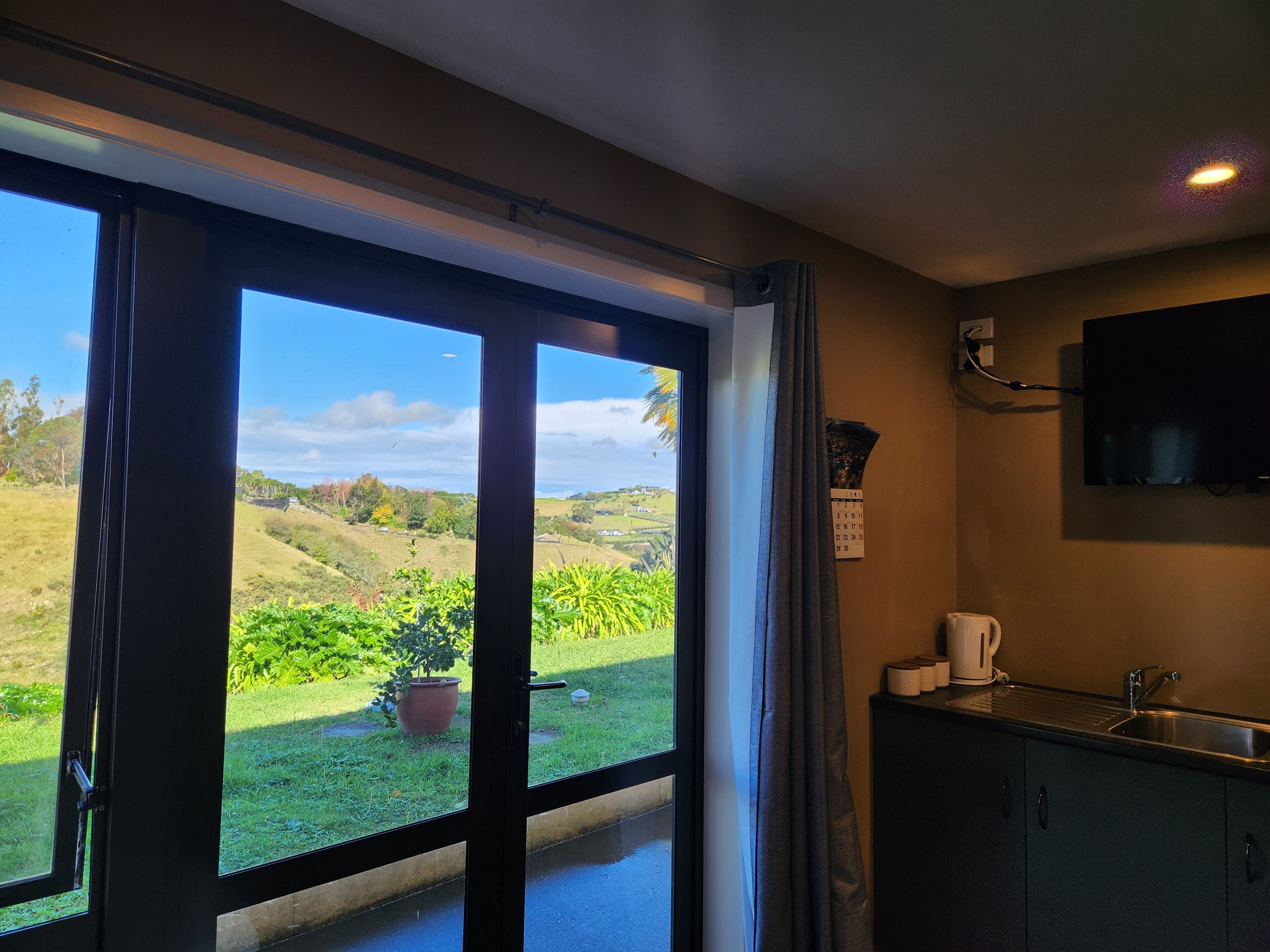 A room with a large sliding glass door leading to a garden, a kitchen area with a sink and cabinets, and a flat-screen TV mounted on the wall.
