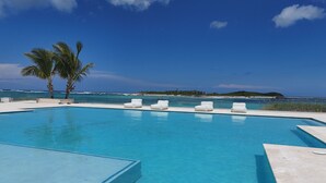 Una piscina azul cristalina con escalones de acceso, rodeada de sillas reclinables blancas y palmeras, con vista al océano y una isla distante.