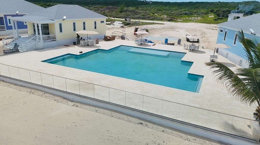 Une piscine rectangulaire dotée d’une terrasse blanche, entourée de chaises longues et de parasols, avec des bâtiments visibles à l’arrière-plan.