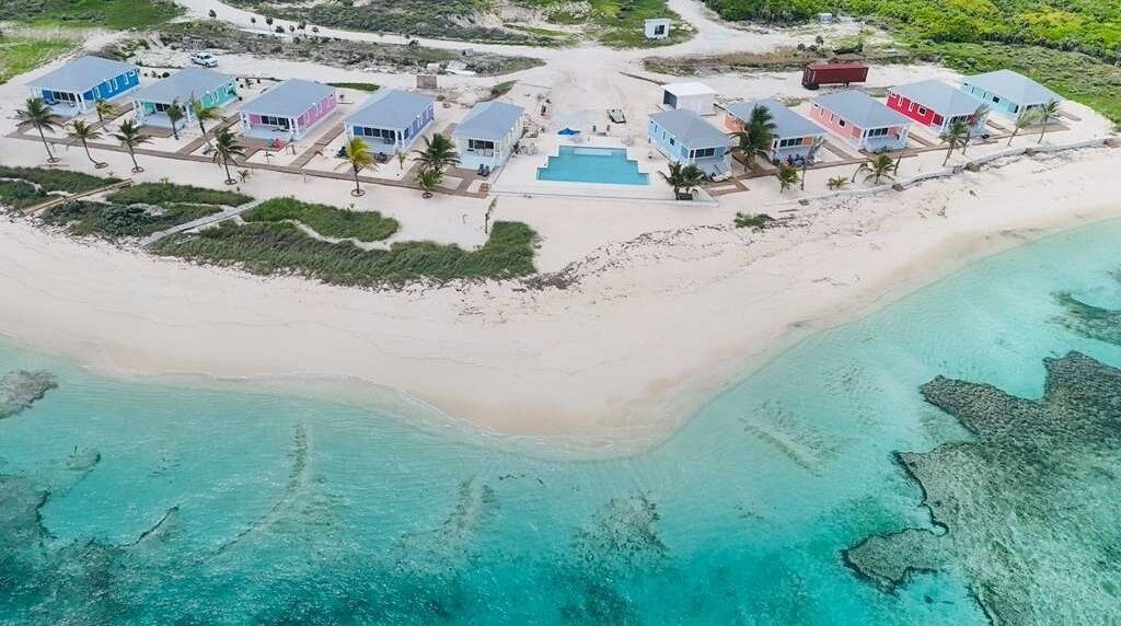 Vista aérea de un resort frente a la playa con varias edificaciones, una piscina y aguas turquesas cristalinas.