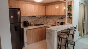 Eine moderne Küche mit Holzschränken, einer Marmorarbeitsplatte, einem eingebauten Gasherd und einem Kühlschrank. Zwei Barhocker und eine kleine Kochinsel mit Spüle ergänzen das Bild.