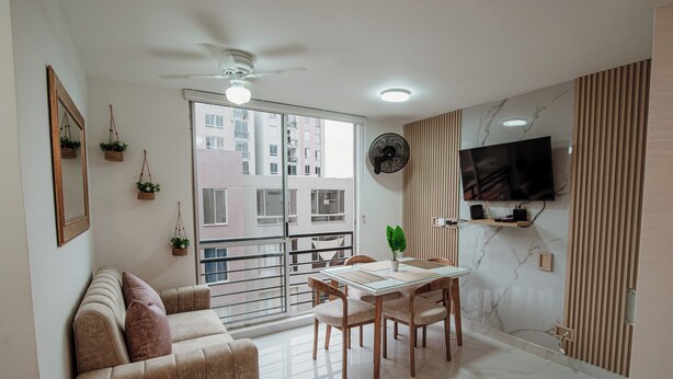 Ein Wohnzimmer mit einer Couch, einem Esstisch, Stühlen, einem Fernseher und einem Balkon mit Blick auf Gebäude.