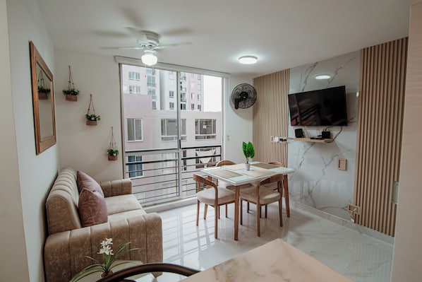 A living room with a sofa, dining table, chairs, a TV, and a balcony with a view of buildings.