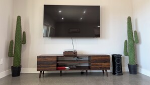 A living room with a flat-screen TV mounted on the wall, a wooden entertainment center, and two potted cacti. - 2-bedroom house in charming Joshua Tree with pool (Joshua Tree)
