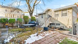 Un patio trasero residencial con terraza, un espacio cubierto para sentarse, una escalera que conduce a un segundo piso y un árbol.