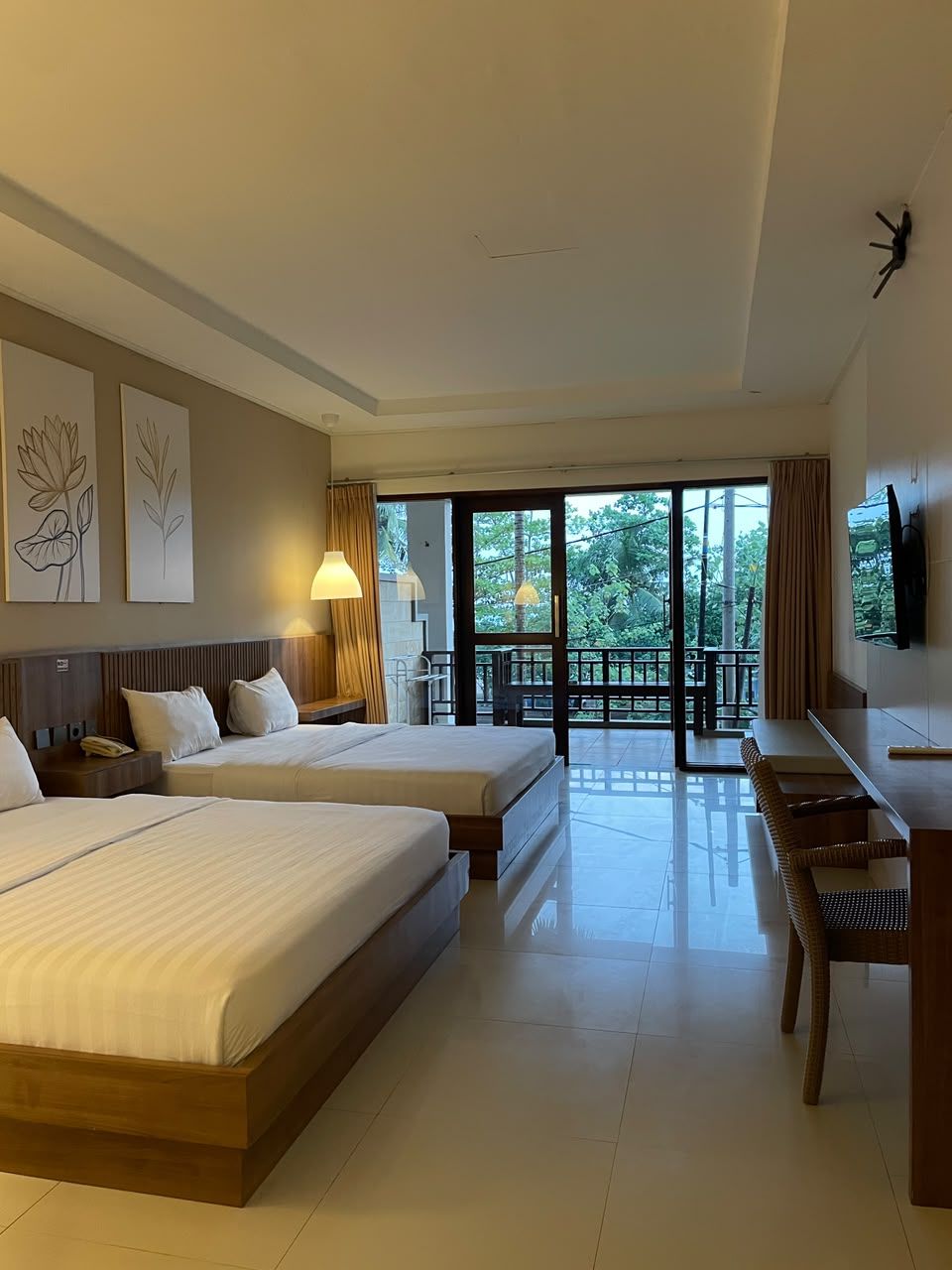 Habitación de hotel con dos camas, un escritorio, una silla, un televisor y un balcón con vistas a árboles.