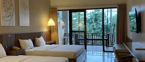 A hotel room with two beds, a desk, a chair, a TV, and a balcony with a view of trees.