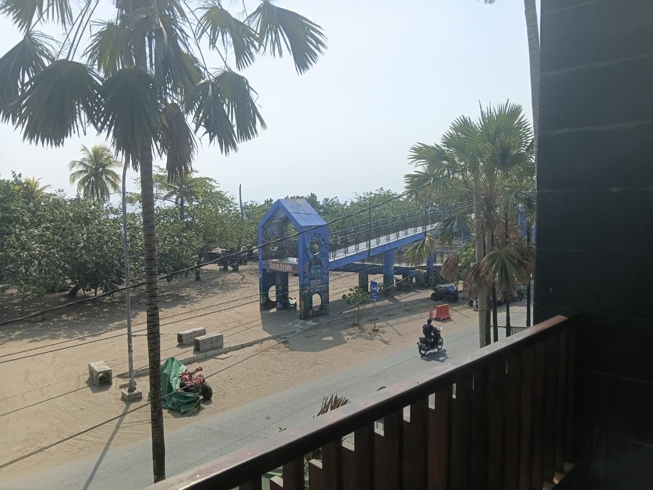 Vista desde un balcón sobre una calle con un puente peatonal azul, palmeras y una motocicleta.