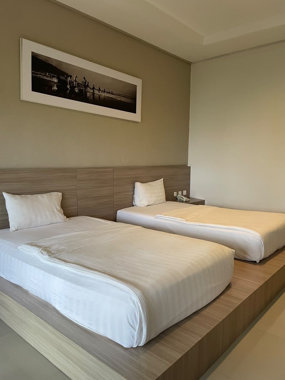 Habitación de hotel con dos camas, cabecero, mesita de noche con teléfono y un cuadro en la pared.