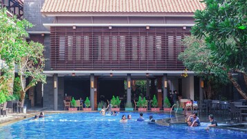 Zona de piscina con varias personas, un área de estar cubierta y un edificio con tejado de baldosas.