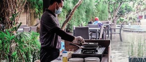 A person cooking outdoors with a table, chairs, and a water feature in the background.
