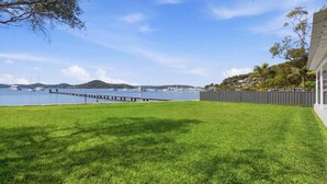A grassy yard with a dock extending into a body of water, surrounded by hills and a fence. - Central Coast Waterfront | 2BR + Jetty Access (Koolewong)