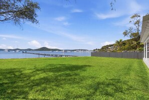 A grassy yard with a dock extending into a body of water, surrounded by hills and a fence. - Central Coast Waterfront | 2BR + Jetty Access (Koolewong)