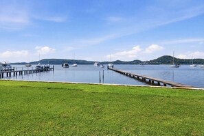 A marina with boats docked, a grassy area in the foreground, and hills in the background. - Central Coast Waterfront | 2BR + Jetty Access (Koolewong)