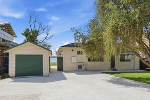 A row of single-story houses with attached garages, a clear sky, and a tree with red flowers. - Central Coast Waterfront | 2BR + Jetty Access (Koolewong)