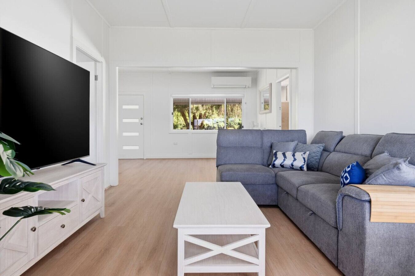 A modern living room with a grey sofa, a flat-screen TV, a white coffee table, and a view of a garden through a window.