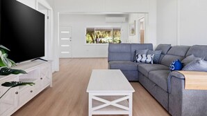 A modern living room with a grey sofa, a flat-screen TV, a white coffee table, and a view of a garden through a window. - Central Coast Waterfront | 2BR + Jetty Access (Koolewong)