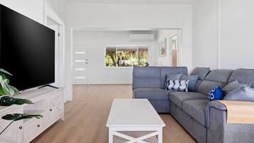 A modern living room with a grey sofa, a flat-screen TV, a white coffee table, and a view of a garden through a window.