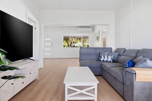 A modern living room with a grey sofa, a flat-screen TV, a white coffee table, and a view of a garden through a window. - Central Coast Waterfront | 2BR + Jetty Access (Koolewong)