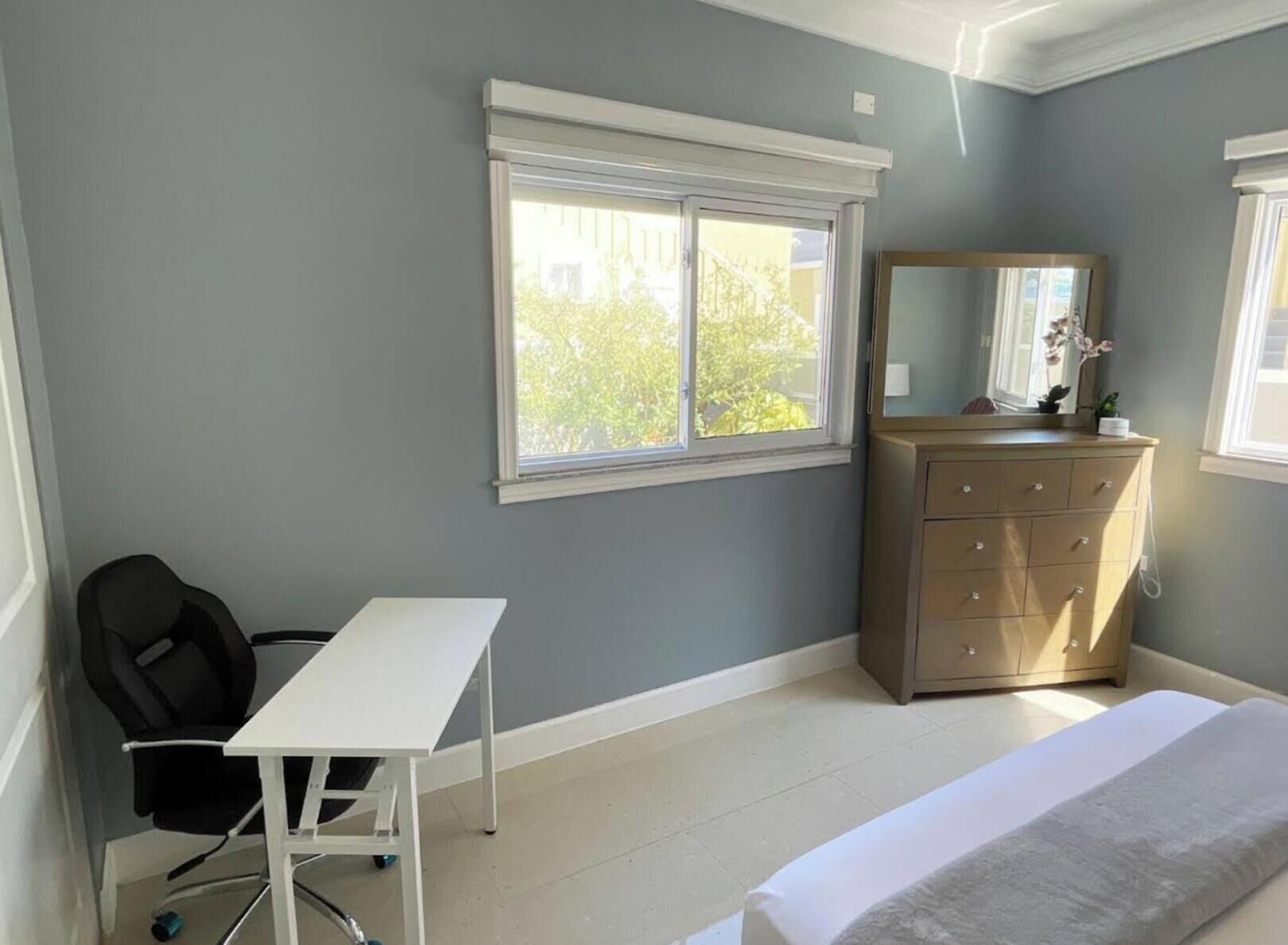 A bedroom with a desk, chair, dresser, and a large window.