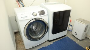 Een wasruimte met een Samsung wasmachine en droger, een blauwe mat en een witte muur met stopcontacten.