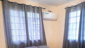 Un lit simple avec une couvre-lit grise, une chaise blanche et une fenêtre avec des rideaux gris.