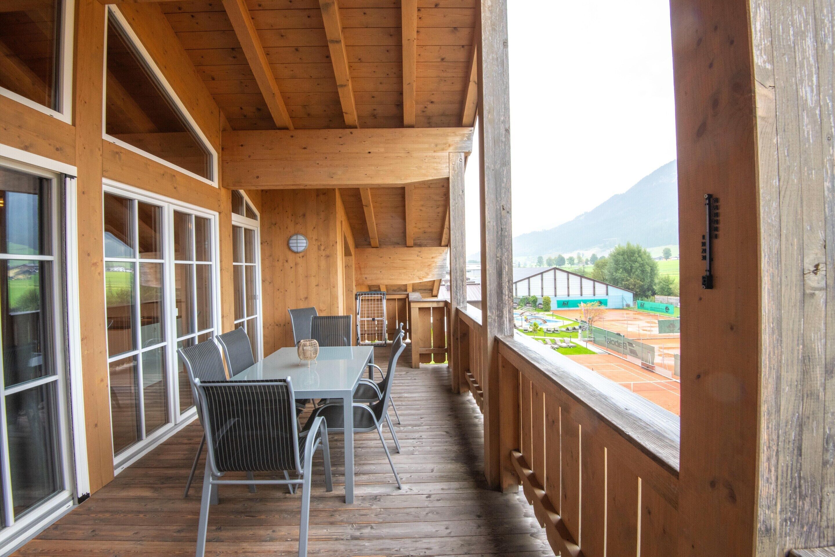 A wooden deck with a table and chairs, large windows, and a view of a tennis court and mountains.