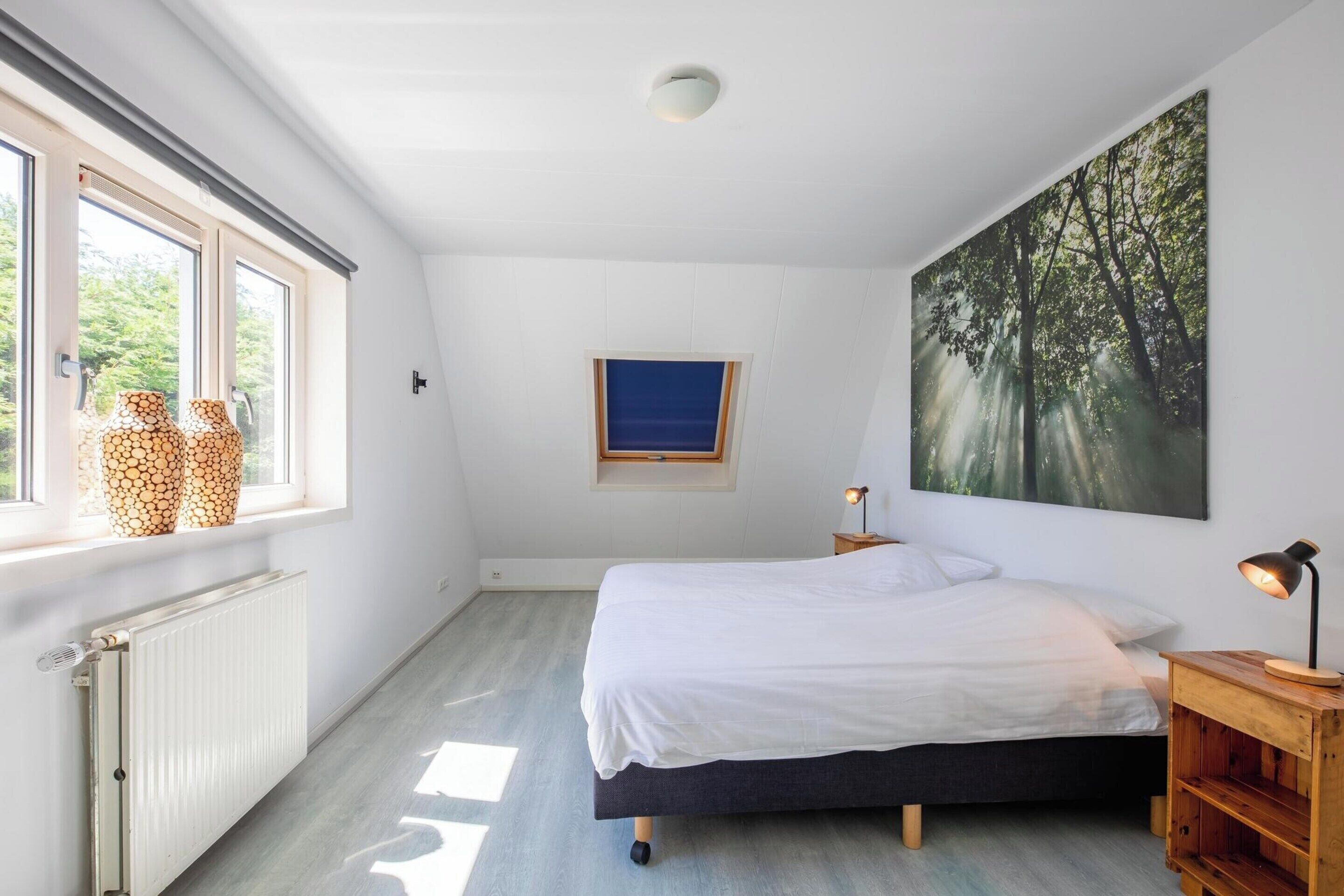 Un dormitorio con una cama, una ventana con jarrones, un radiador, un estante en la pared, un cuadro y una mesita de noche con una lámpara.