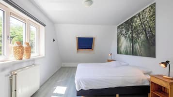 Un dormitorio con una cama, una ventana con jarrones, un radiador, un estante en la pared, un cuadro y una mesita de noche con una lámpara.