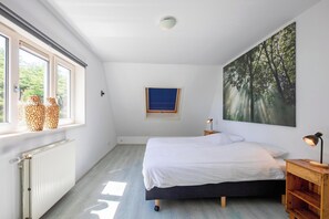 Un dormitorio con una cama, una ventana con jarrones, un radiador, un estante en la pared, un cuadro y una mesita de noche con una lámpara.