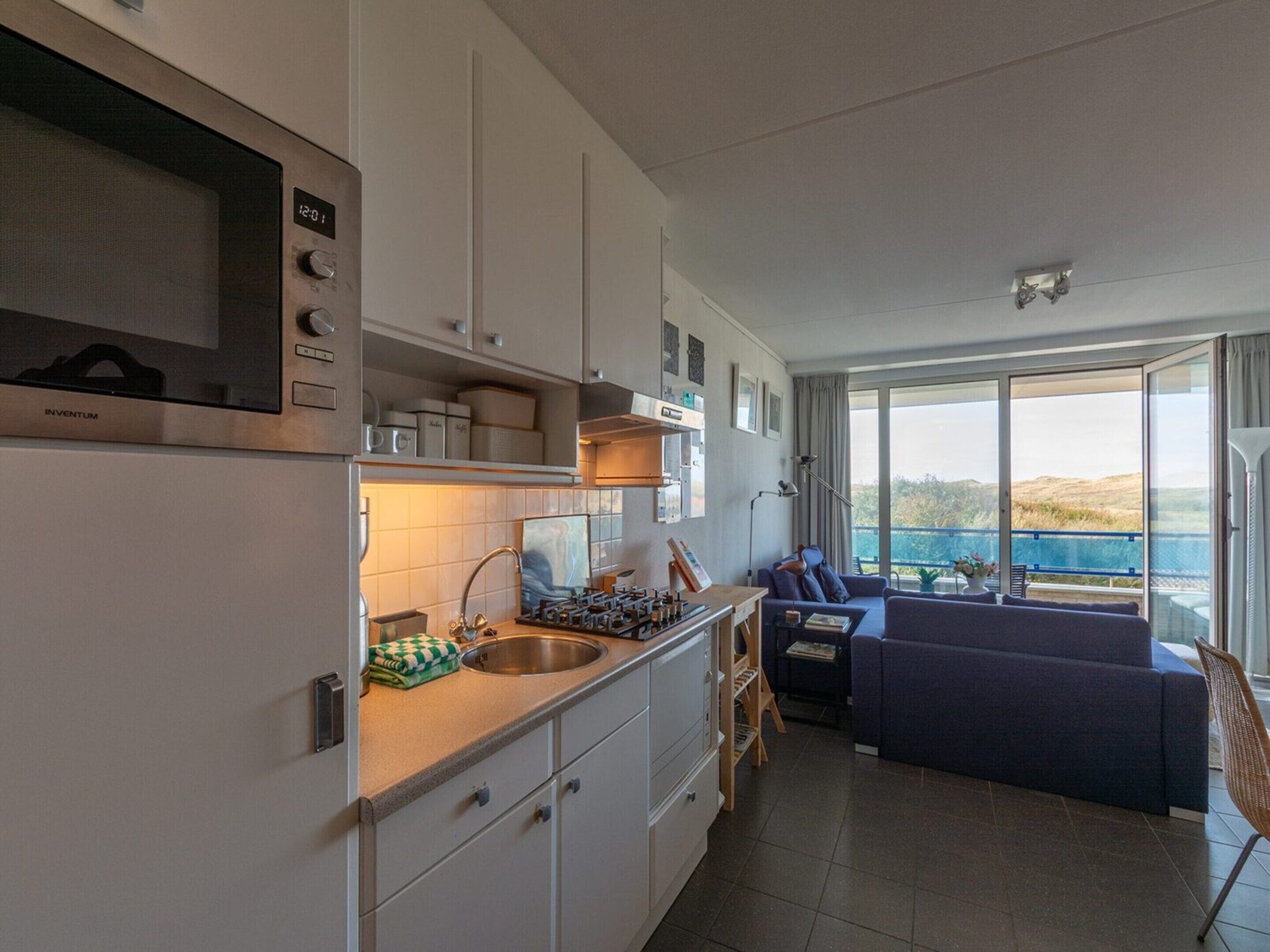A modern kitchen with white cabinets, a stainless steel sink, and a microwave. There is a dining area with a blue sofa and a small table. The room has large windows offering a view of the outdoors.