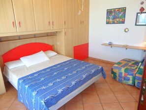 Un dormitorio con cama, cabecera roja, colcha azul, aparador rojo y una estantería con un cuadro.