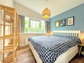A bedroom with a bed, a wooden crib, a window with curtains, and a hanging light fixture. - Bungalow in Sint Maartenszee With Garden Views (Sint Maartensvlotbrug)