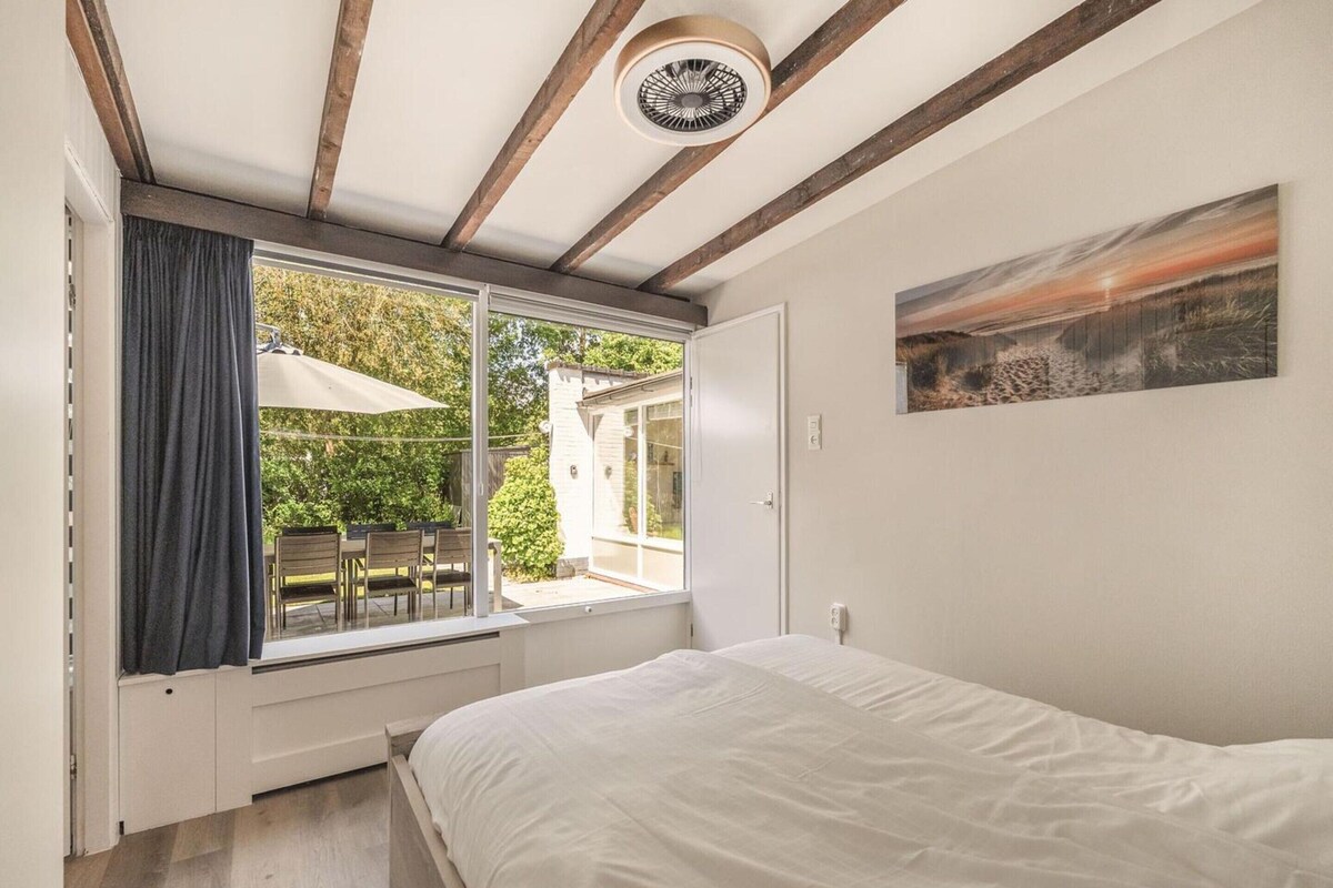 A bedroom with a bed, a window with curtains, a ceiling fan, and a painting on the wall.