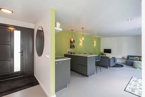 A modern kitchen with a built-in island, a dining area with a gray sofa, and a living room with a TV. - Bungalow in Sint Maartenszee With Garden Views (Sint Maartensvlotbrug)
