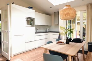A modern kitchen with white cabinets, a wooden dining table, and a hanging light fixture. - Bungalow in Sint Maartenszee With Garden Views (Sint Maartensvlotbrug)