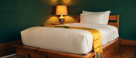 Uma cama de madeira com roupa de cama branca e um cobertor amarelo, um tapete estampado e uma luminária em uma mesa de cabeceira.