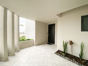 Un couloir au sol carrelé de carreaux blancs, une porte noire et un petit espace jardin avec des plantes.