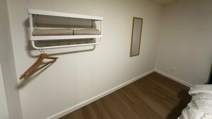 Une chambre avec un plancher en bois, un lit avec une literie blanche, une étagère blanche fixée au mur avec une serviette, et un miroir.