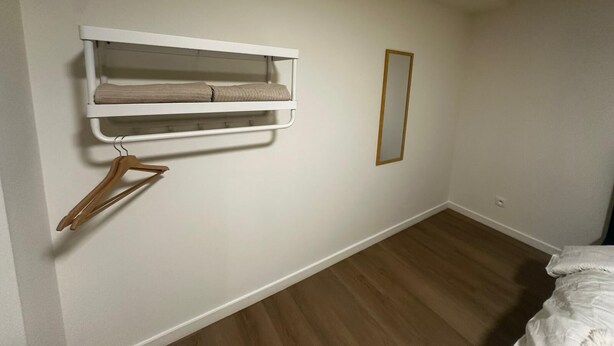 Una camera da letto con pavimento in legno, un letto con biancheria bianca, uno scaffale a parete bianco con un asciugamano e uno specchio.