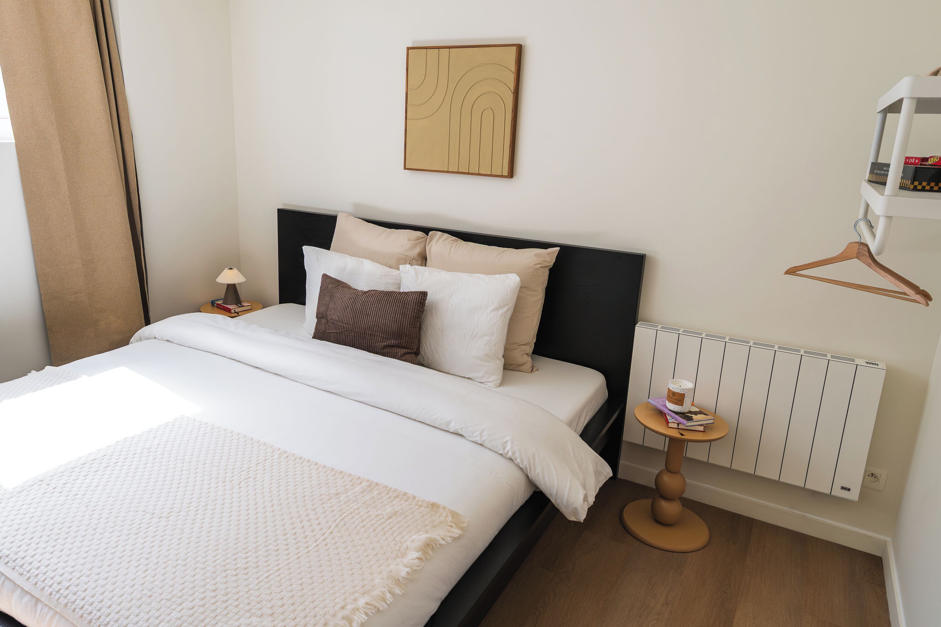 A neatly made bed with a white comforter and pillows, a bedside table with a lamp, a wall-mounted radiator, and a small wooden table with a cup and book.