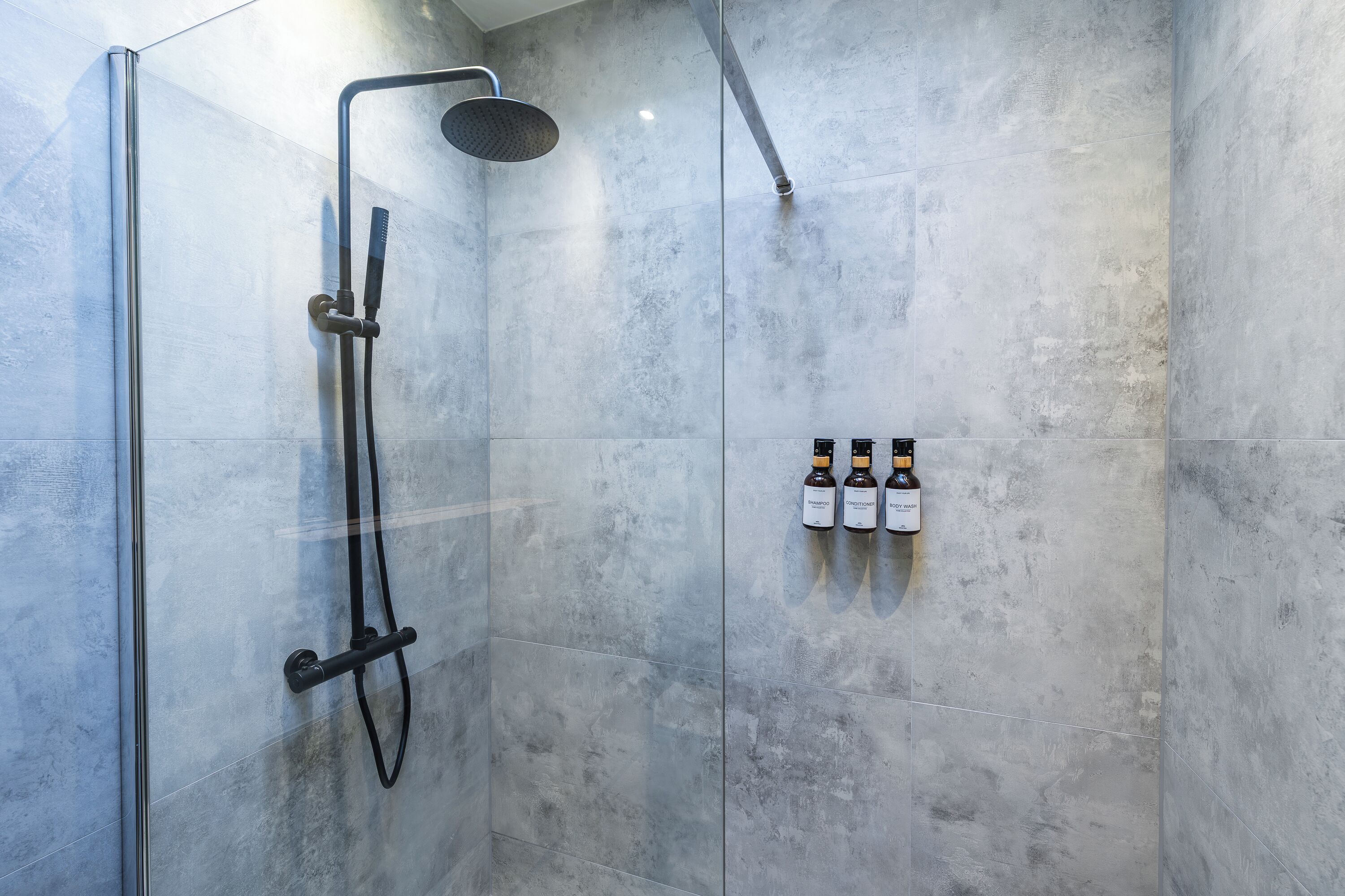 A shower area with a glass door, a handheld showerhead, and three bottles of shampoo on the wall.